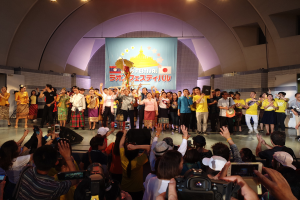 Gruppe von Menschen auf einer Bühne mit Kameras, Handys und Musikinstrumenten während einer Festivaleröffnung mit Bannern, Wänden und Deckenlampen im Hintergrund.