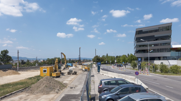 Baustelle mit parkenden Autos, Gebäuden, Straßeninfrastruktur, Vegetation, Sand, Kränen, Bahnschienen, Stromleitungen, fernen Hügeln und bewölktem Himmel.