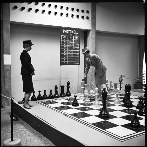 Zwei Frauen spielen Schach auf einem großen schwarzen und weißen Brett vor einer Wand mit einem Brett daran, mit Schachfiguren darauf angeordnet.