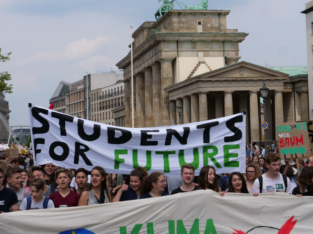 Eine Gruppe von Schülern marschiert in Berlin, die eine bunt bemalte Plakette mit der Aufschrift "Schüler für die Zukunft" trägt, mit Gebäuden, Bäumen und Himmel im Hintergrund.
