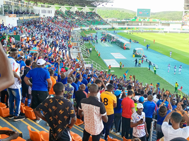 Eine große Menschenmenge versammelt sich vor einem eingezäunten Fußballfeld, wobei einige auf Stühlen sitzen und andere stehen, während Hügel und ein klarer blauer Himmel die Kulisse bilden.