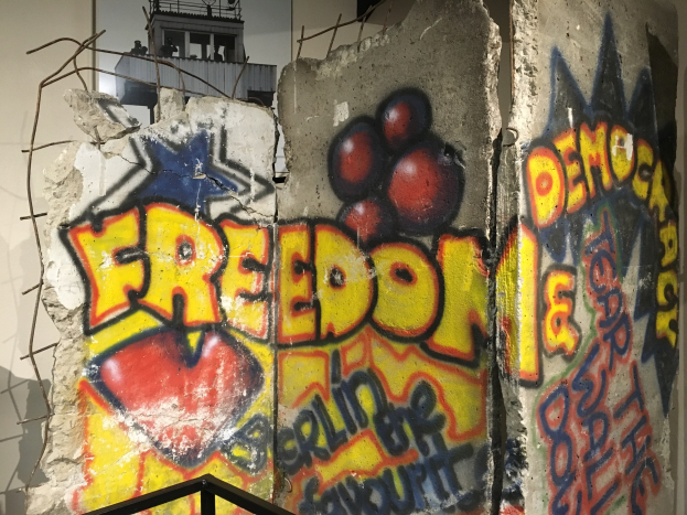 Mauer mit Graffiti, das in fetten schwarzen Buchstaben "Freiheit und Demokratie" liest, ein Geländer unten und ein Gebäude mit Fenstern im Hintergrund.