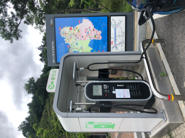 Eine Ladesäule für Elektrofahrzeuge am Straßenrand mit einem Motorrad in der Nähe, Bäumen und einem klaren blauen Himmel im Hintergrund und einem digitalen Anzeigeboard, das anzeigt, dass es eine E-Bike-Ladestation ist.