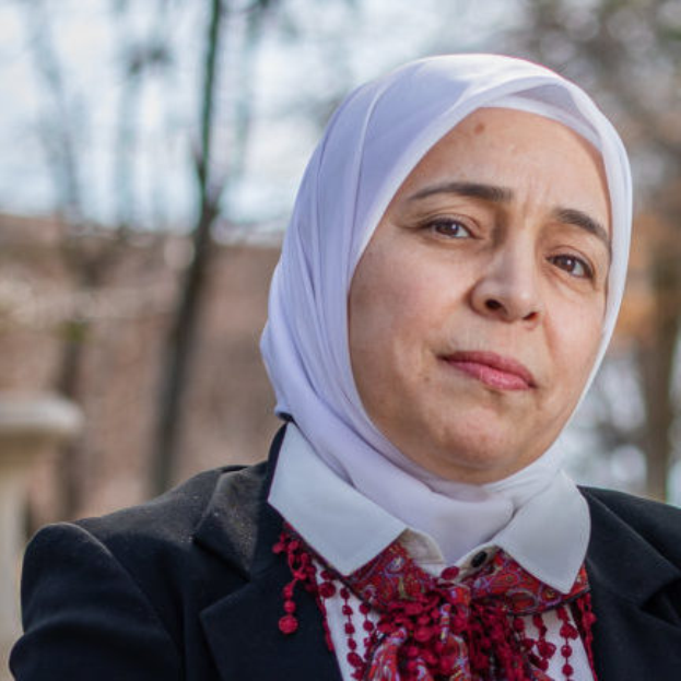 Juli Tymoshenko, eine Frau mit einem weißen Kopftuch und einer roten Schärpe, steht vor einem unscharfen Hintergrund aus Bäumen und einem Gebäude.