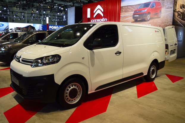 Ein weißer Citroën Berlingo Van wird auf einer Autauschau präsentiert, umgeben von schwarzen Schlafzimmermöbeln, während die Wände mit Brettern und Lichtern geschmückt sind.