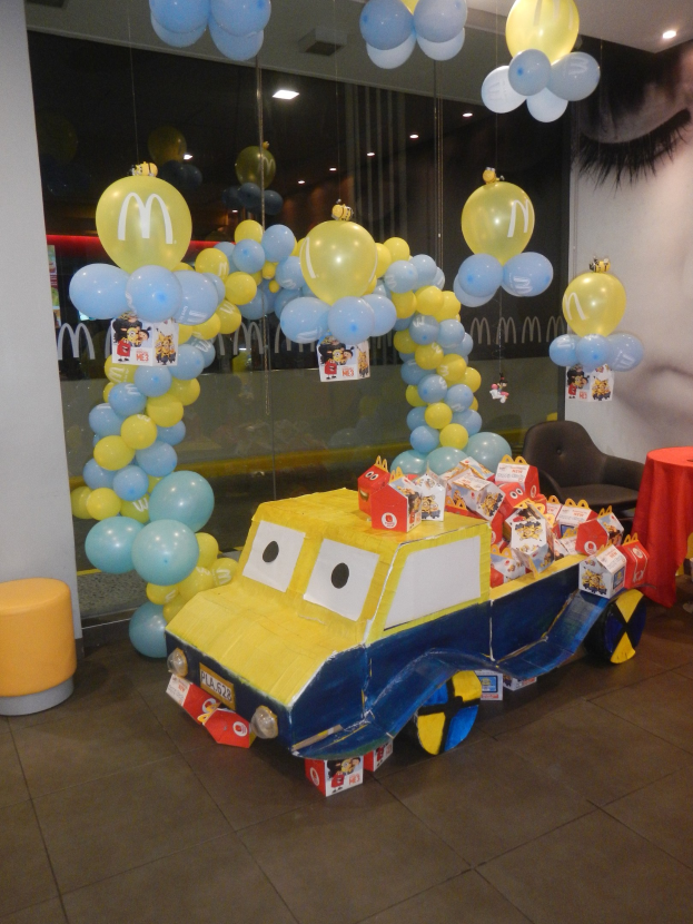 Eine festliche McDonald's-Geburtstagsparty-Szene mit einem gelben und blauen Lastwagen, der mit Ballons und anderen Dekorationen geschmückt ist, einem Tisch mit rotem Tuch und Gegenständen, Hockern, einer Glaswand und Deckenleuchten.