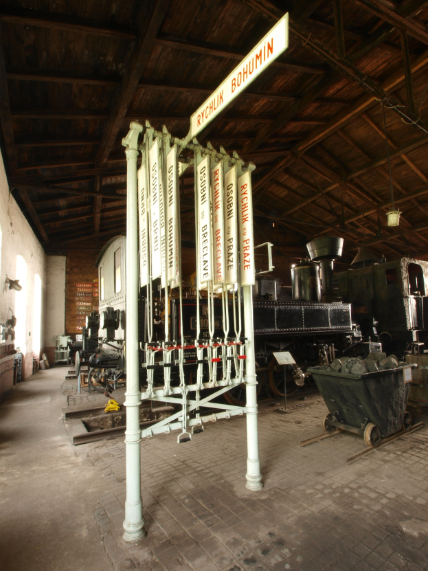 Ein Bahnhof mit einem Zug auf den Gleisen, einem Handwagen auf dem Boden, einem Pfahl mit einer Tafel, einer Wand auf der linken Seite, einer Decke oben und einer Signalbox in der Mitte.