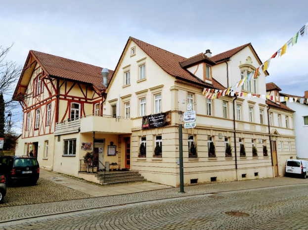Großes Gebäude namens Stadtbahnhof in Baden-Württemberg mit vielen Fenstern, umgeben von Bäumen, Straßenlaternen, Fahrzeugen, Fußgängern, Werbetafeln und dekorativen Fahnen unter einem bewölkten Himmel.
