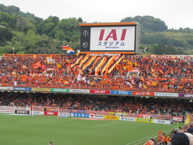 Ein Fußballspiel in einem Stadion mit großer Zuschauermenge, saftigem Grün, einem Tor, Bannern, Fahnen, einem großen Bildschirm, Bäumen und einem klaren blauen Himmel.