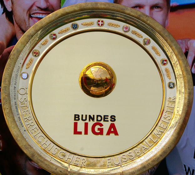 Goldener Schild mit 'Bundesliga'-Text in den Händen eines Mannes, im Hintergrund ein Banner mit drei Personen.