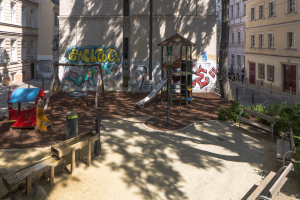 Spielplatz mit Graffiti in einer städtischen Umgebung, umgeben von Gebäuden, Bäumen, Bänken und einem Mülleimer, mit Menschen und Fahrzeugen auf der Straße und einem sichtbaren Himmel im Hintergrund.