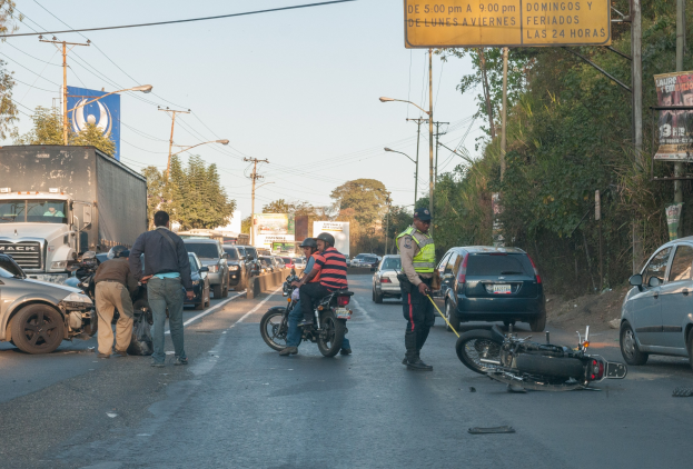 Gruppe von Menschen um ein verunglütztes Motorrad am Straßenrand mit mehreren Fahrzeugen, darunter ein Lastwagen, im Hintergrund und Bäumen, Masten, Lichtern und Schildern an der Straße.