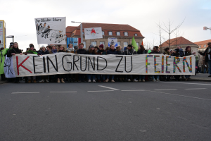 Demonstranten mit einem Transparent mit der Aufschrift 'Kein Grund zur Feier' gegen deutsche Sparmaßnahmen, im Hintergrund Gebäude, Bäume und einen klaren Himmel.