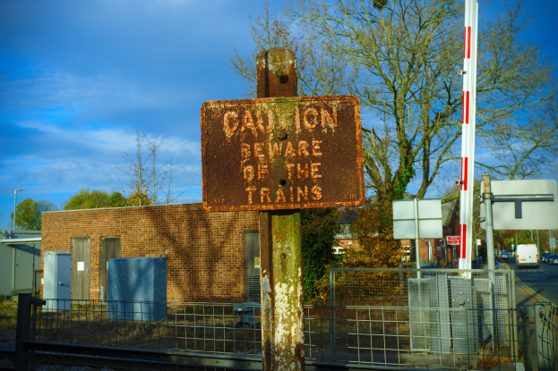 Warnschild an einer Bahnschiene umgeben von einem Metallzaun, Bäumen, Strommasten, einem Gebäude mit Fenstern, Containern, Fahrzeugen auf einer Straße und einem bewölkten Himmel.