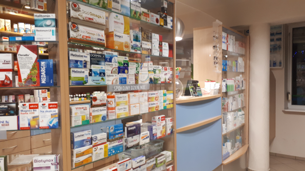 Eine Apotheken-Inneneinrichtung mit Regalen, die mit verschiedenen Medikamenten bestückt sind, einer zentralen Säule und einem Glasfenster auf der rechten Seite, mit der Decke oben.
