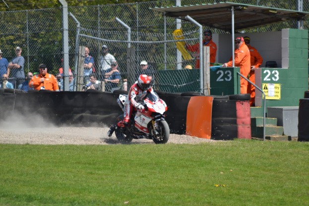 Eine Person in einem Helm und Handschuhen fährt ein Motorrad über ein grasbewachsenes Feld, mit Zuschauern in Mützen und Schutzbrillen, einem Zaun, Stufen und einem nummerierten Schuppen im Hintergrund.