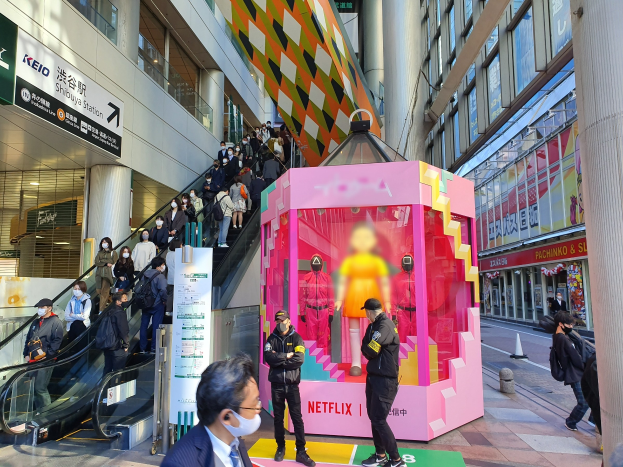 Eine Gruppe von Menschen steht vor einem Gebäude mit Glaswänden und Texttafeln, einige tragen Masken, während eine Rolltreppe mit Fahrgästen zu sehen ist; eine pinkfarbene Box in der Mitte zeigt Schaufensterpuppen und trägt den Text "Netflix's new Netflix booth in Tokyo."