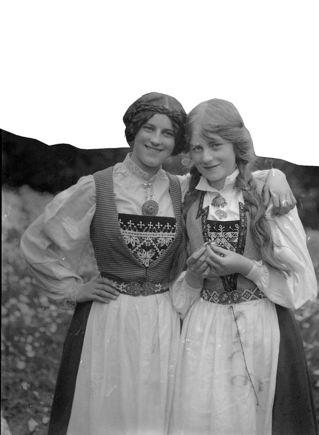 Zwei junge Frauen in traditioneller bayrischer Tracht stehen lachend, sie tragen weiße und schwarze bestickte Kleider vor einem Hintergrund aus grünen Pflanzen und einem hellen Himmel.