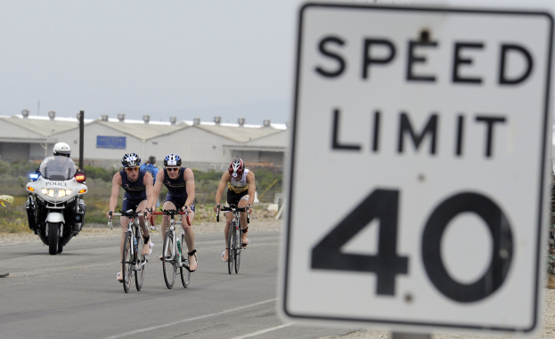 Eine Gruppe von Menschen mit Helmen fährt Fahrräder auf einer Straße neben einem Geschwindigkeitsbegrenzungsschild, mit Gebäuden, Masten, Pflanzen, Bäumen und einem klaren blauen Himmel im Hintergrund.