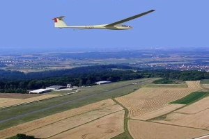 Segelflugzeug über einem Feld mit Bäumen und Gebäuden im Hintergrund, mit Text oben im Bild.