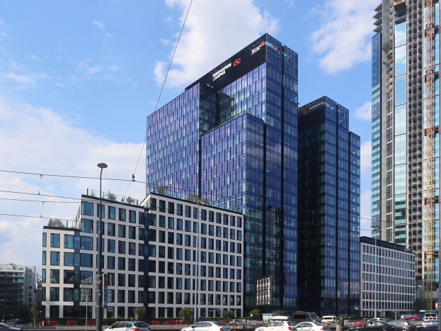 Großes modernes Gebäude mit zahlreichen Fenstern, identifiziert als Deutsche Bank Hauptquartier in Frankfurt, Deutschland, umgeben von städtischer Infrastruktur einschließlich Straßenlaternen, Straßenlaternen, elektrischen Polen, elektrischen Kabeln, Kraftfahrzeugen auf der Straße, Büschen, Bäumen und einem bewölkten Himmel.