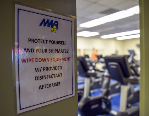 Zeichen an einer Turnhallentür mit der Aufschrift "Schütze dich und deine Schiffskameraden" und Laufbänder sowie Deckenleuchten im Hintergrund.