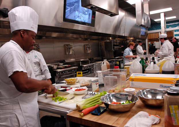 Eine Gruppe von Köchen in einer Küche, die auf einem Schneidbrett Lebensmittel zubereiten, mit einem Koch im Vordergrund in einer weißen Schürze und Mütze, der ein Messer hält, und umgeben von verschiedenen Utensilien, Schüsseln, Gemüse, einem Handy, Flaschen und anderen Gegenständen, mit einem Fernseher und Deckenleuchten im Hintergrund.