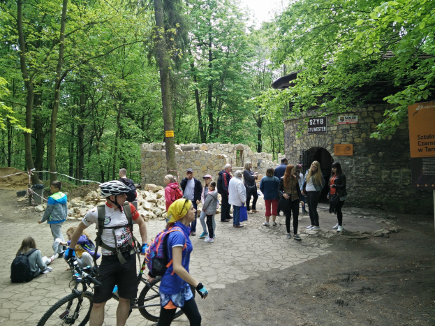 Gruppe von Menschen, die einen holprigen Pfad in einem Waldgebiet entlanggehen, einige tragen Helme und haben Taschen oder Fahrräder dabei, im Hintergrund eine Steinmauer und ein Schild, das den Eingang zum Szyb Stylweste Mountainbike Trail anzeigt.