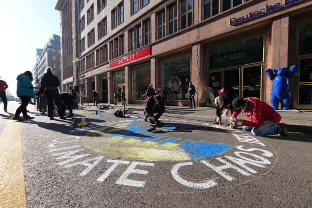 Menschen sitzen vor einem Gebäude mit Fenstern und Namensschildern auf dem Boden, umgeben von Flaschen und anderen Gegenständen während einer Klimaprotest in Berlin.