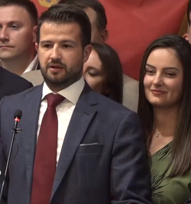 Ein Mann im Anzug und Krawatte steht an einem Podium mit einem Mikrofon und spricht zu einer Menge, mit einem Banner im Hintergrund.