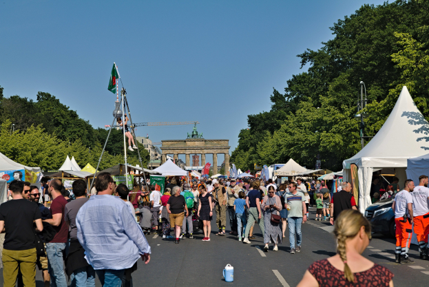 Eine Menschenmenge geht eine Straße entlang, die mit Zelten, Fahrzeugen und Bäumen gesäumt ist, mit einem Bogen und einem klaren blauen Himmel im Hintergrund und Polen mit Fahnen auf der linken Seite, die likely das Oktoberfest in München, Deutschland, darstellen.