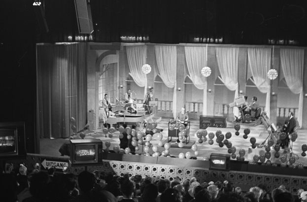 Schwarzes und weißes Bild von Menschen auf einer Bühne mit Ballons und Fernsehern im Hintergrund.