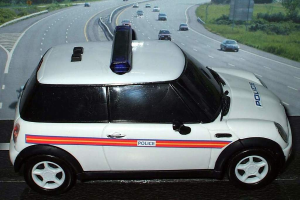 Ein weißes Polizeiauto ist im Vordergrund geparkt, mit vielen Autos, die sich dahinter auf der Straße bewegen, umgeben von Gras und Bäumen auf beiden Seiten.