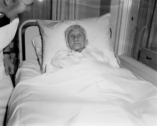 Alte Frau im Krankenhausbett mit einer Krankenschwester daneben, Bettdecke und Kissen weiß, Fenster mit Vorhängen im Hintergrund.