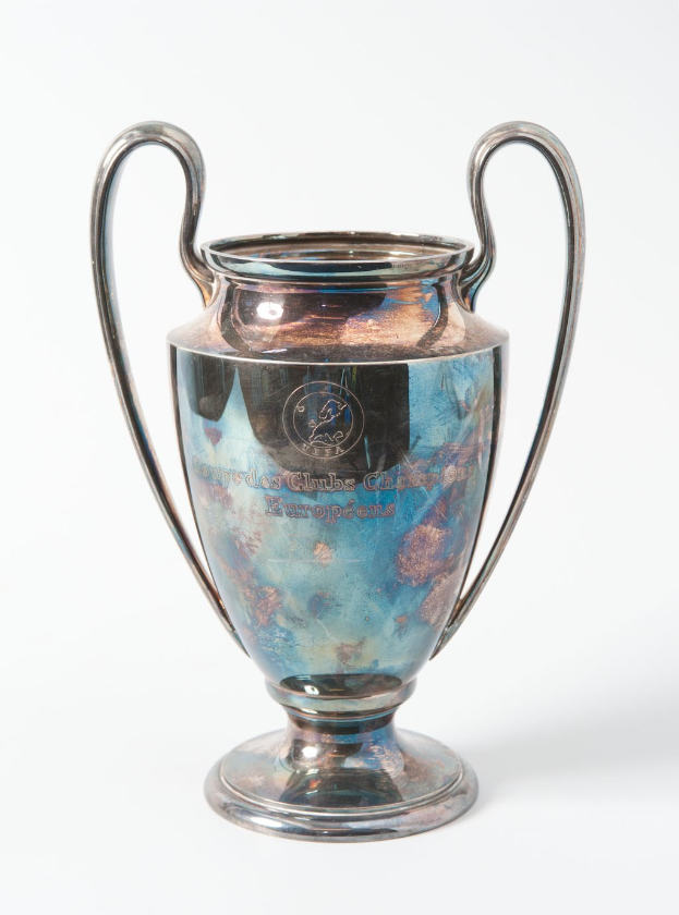 Der UEFA Champions League Pokal, ein silberner und blauer Stahlbecher mit zwei Griffen, der mit "UEFA Champions League" beschriftet ist, vor einem weißen Hintergrund.