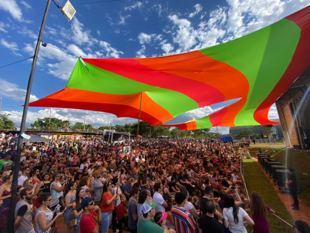 Eine große Menschenmenge steht vor einer Regenbogenflagge mit einem Gebäude, Lautsprechern auf der Bühne, Bäumen und Wolken im Hintergrund während des 10-jährigen Jubiläums eines Festivals.