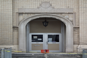 Ein Eingang eines Backsteingebäudes mit gelbem Absperrband, Treppen mit Maschendrahtzaun, eine Tür mit Papieren, eine hängende Lampe und eine Wandinschrift, die wahrscheinlich den Namen der Schule angibt.