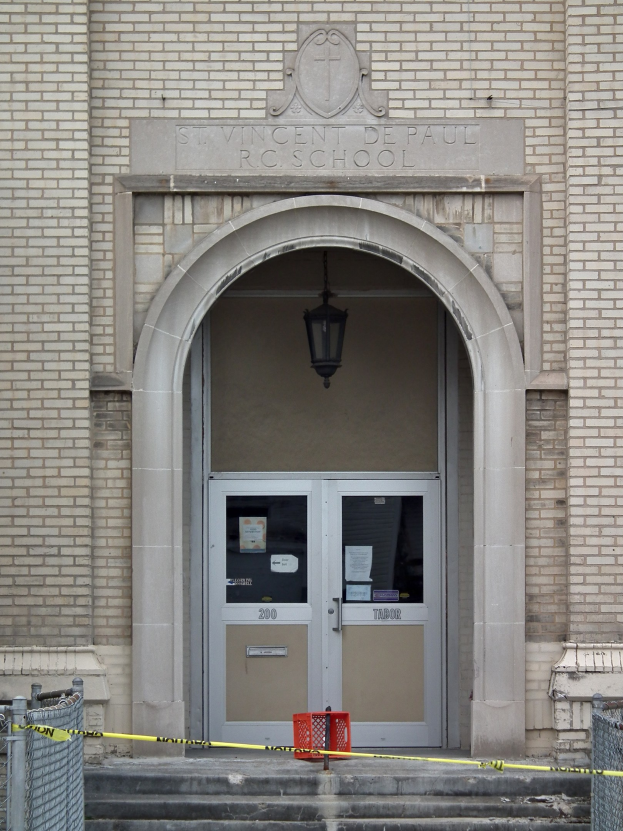 Ein Eingang eines Backsteingebäudes mit gelbem Absperrband, Treppen mit Maschendrahtzaun, eine Tür mit Papieren, eine hängende Lampe und eine Wandinschrift, die wahrscheinlich den Namen der Schule angibt.