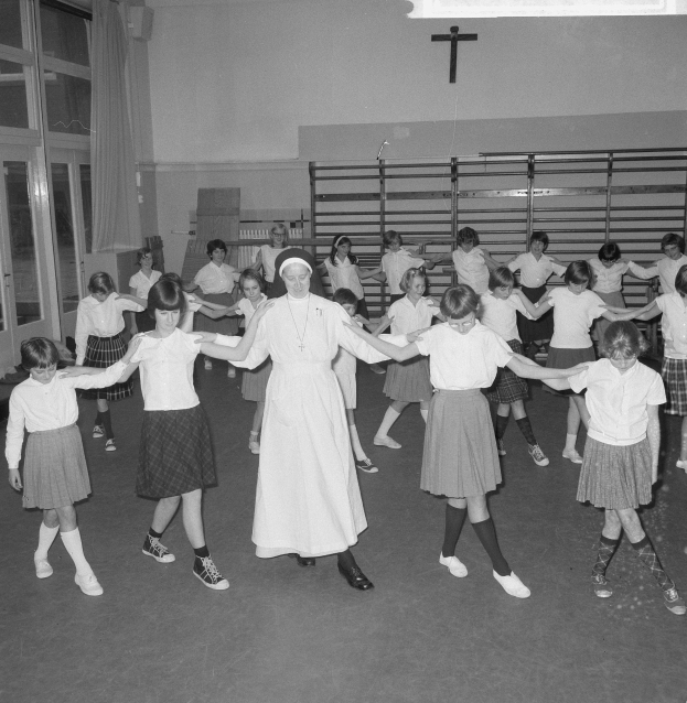 Schwarzes und weißes Bild von Kindern in Schuluniformen, die in einem Raum mit einem Kreuzsymbol an der Wand, Glastüren mit Vorhängen und Deckenleuchten tanzen.