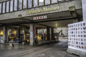 Außenansicht des Deutschen Museums in Berlin mit einem großen Glasgebäude, Säulen, einem Namensschild, einem Anzeigebrett mit Bildern und Text auf der rechten Seite und umgeben von anderen Gebäuden, Bäumen und Himmel.
