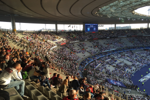 Große Menschenmenge in einem Stadion bei einem Fußballspiel, mit einer Bühne rechts, Fahnen, Stangen, einem Bildschirm und dem Allianz Arena in München im Hintergrund.