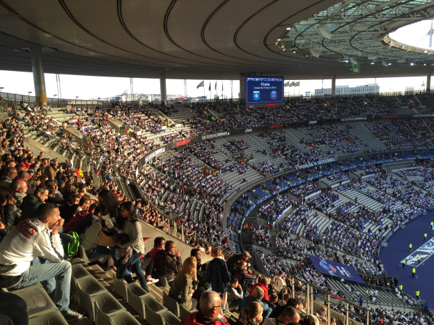 Große Menschenmenge in einem Stadion bei einem Fußballspiel, mit einer Bühne rechts, Fahnen, Stangen, einem Bildschirm und dem Allianz Arena in München im Hintergrund.