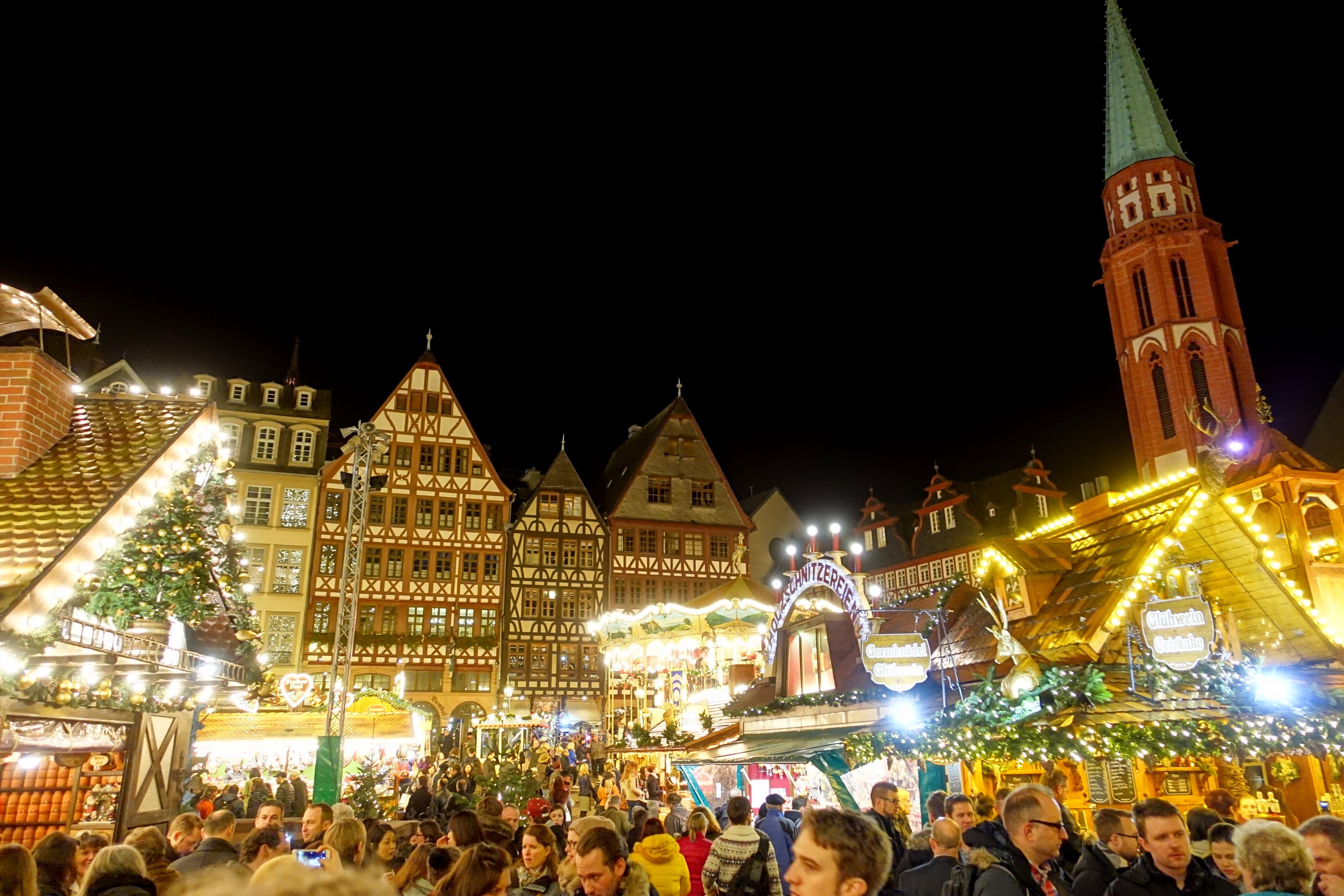 Ein geschäftiger Weihnachtsmarkt in Nürnberg, Deutschland mit Menschen um dekorierte Stände, festliche Lichter, Gebäude mit Fenstern, ein Uhrenturm und einen dunklen Himmel.