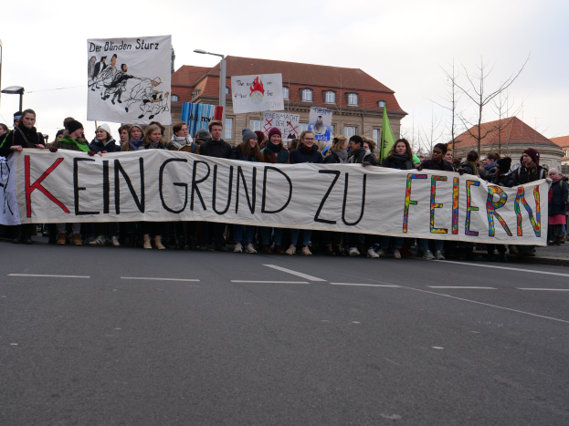 Demonstranten mit einem Transparent "Kein Grund zu Feiern" gegen deutsche Sparmaßnahmen, im Hintergrund Straßeninfrastruktur und Gebäude.