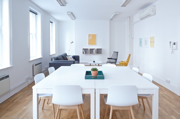 Innenansicht eines Apartments in Berlin mit einem weißen Tisch mit einer Pflanze in einer Schale, Stühlen, einem Sofa mit Kissen, einer Lampe, Regalen mit Gegenständen, einer Klimaanlage, Fotorahmen an der Wand, Fenstern und Deckenleuchten.
