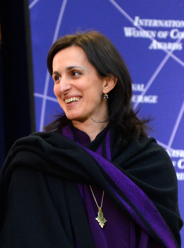 Eine Frau in einem lilafarbenen Kleid und schwarzer Stola steht neben einer anderen Frau, beide lächeln, mit einem Banner im Hintergrund, auf dem "Internationale Frauen der Farbe" steht.