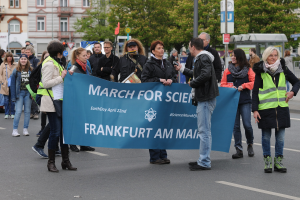 Gruppe von Menschen marschiert auf einer Straße mit einem "March for Science Frankfurt am Main"-Schild, mit Bäumen, Gebäuden und einem klaren Himmel im Hintergrund.