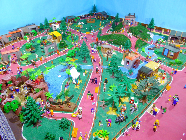 Ein Miniatur-Modell eines Zoos mit Besuchern, umgeben von Bäumen, Häusern, Pflanzen, Gras, einem Zaun und einer Hintergrundwand, mit Text unten.