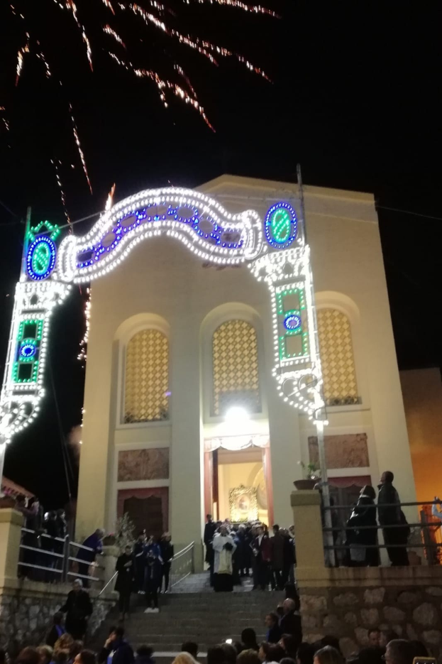 Eine Gruppe von Menschen, die vor einem Gebäude mit einem beleuchteten Bogengang, Treppen mit Geländern und Topfpflanzen steht und Silvester mit einem Feuerwerk im Hintergrund feiert.