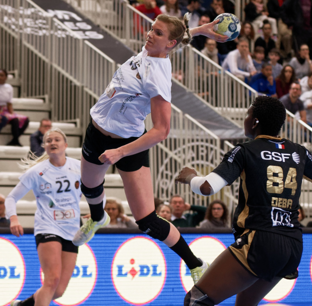 Gruppe von Frauen, die Handball auf einem Platz spielen, mit einem Ball in der Luft, Zuschauern auf Treppen und einem "Handball-Weltmeisterschaft 2019 - Tag 2"-Schild im Hintergrund.